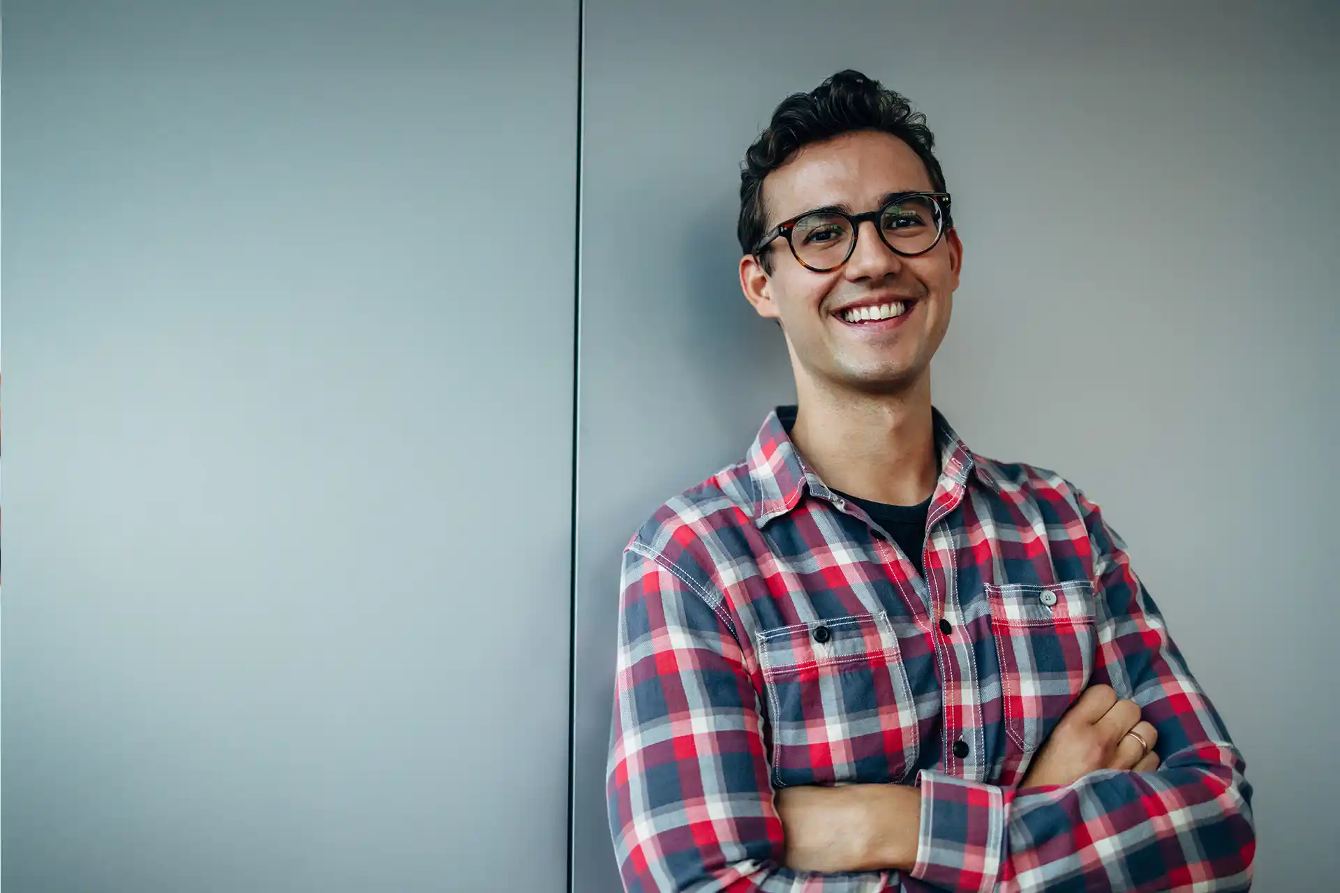 Junger Mann mit Brille und kariertem Hemd steht lächelnd mit verschränkten Armen vor einer Wand – typisches Porträt eines Auszubildenden im kaufmännischen Bereich.