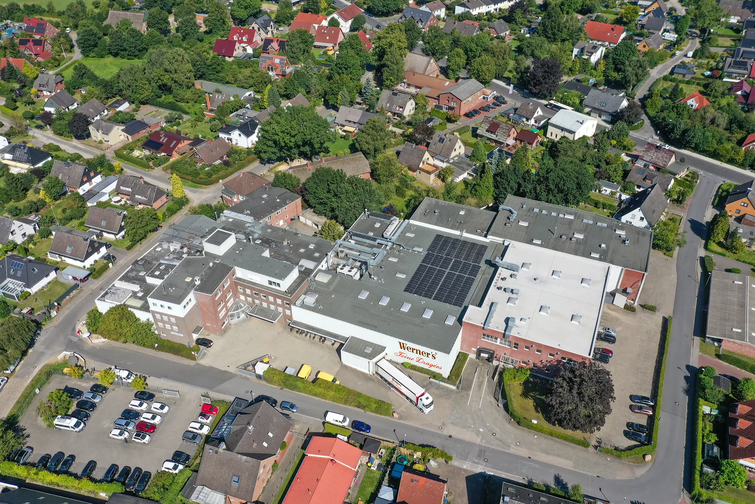 Historic brick building of Werners GmbH & Co. KG with three floors, several windows, and a small canopy over the entrance. The windows on the right side of the building are partially boarded up with wooden planks. In front of the building stands a classic black Mercedes with an open door. The surroundings include a paved street, a low brick wall, and some trees and houses in the background.