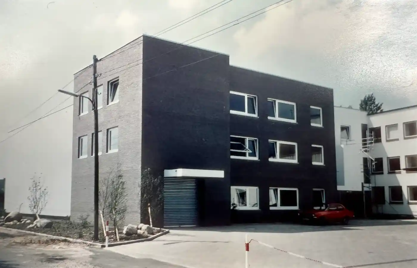 Historic brick building of Werners GmbH & Co. KG with three floors, several windows, and a small canopy over the entrance. The windows on the right side of the building are partially boarded up with wooden planks. In front of the building stands a classic black Mercedes with an open door. The surroundings include a paved street, a low brick wall, and some trees and houses in the background.